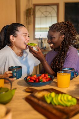 İki kadın mutfak masasında birlikte sağlıklı bir kahvaltı yapıyor, bir anlık şefkat ve kahkaha paylaşıyorlar.