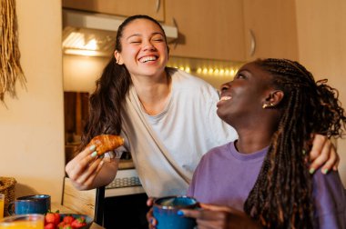 İki kadın kahkahalarını paylaşıyor ve sıcak mutfaklarında birlikte kahvaltı ediyorlar. Sevgi ve bağlarla dolu neşeli bir an yaratıyorlar.