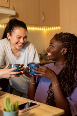 İki kadın kahvaltının tadını çıkarıyor, ellerinde fincanlar ve mutfaklarında neşeli bir anı paylaşıyorlar.