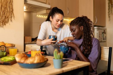 İki kadın mutfak masalarında keyifli bir kahvaltı yapıyorlar, kahve ve sohbetin tadını çıkarıyorlar.