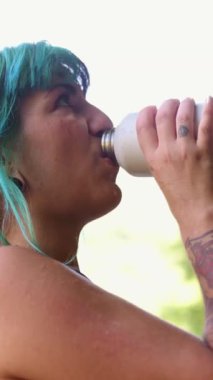 Kantinden su içen turkuaz saçlı susamış bir kadın. Güneşli yeşil bir ormanda yürüyüş yaparken su içen kıza yaklaş.