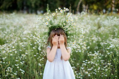 Kafasında çiçek çelengi olan beyaz elbiseli bir kız papatya tarlasında duruyor.