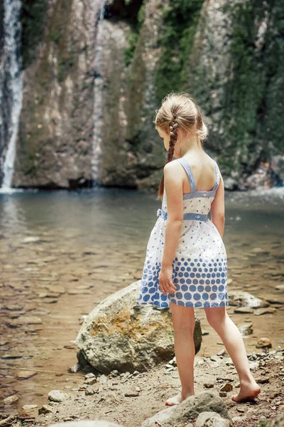 Blondýnka v bílo-modrých šatech stojí na břehu lesní řeky na pozadí vodopádu. — Stock fotografie