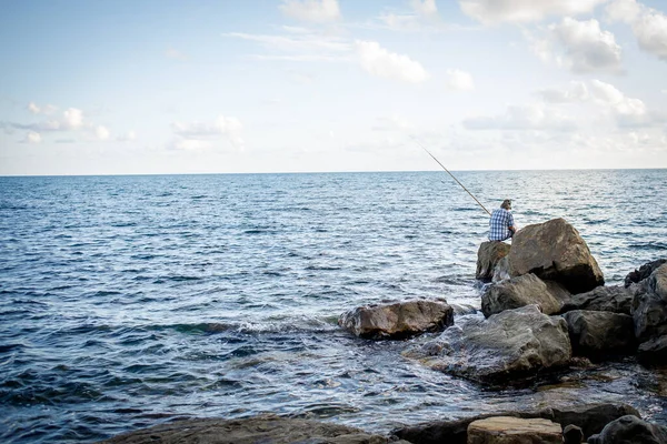 Рыбак рыбачит в море, сидя на больших камнях. — стоковое фото