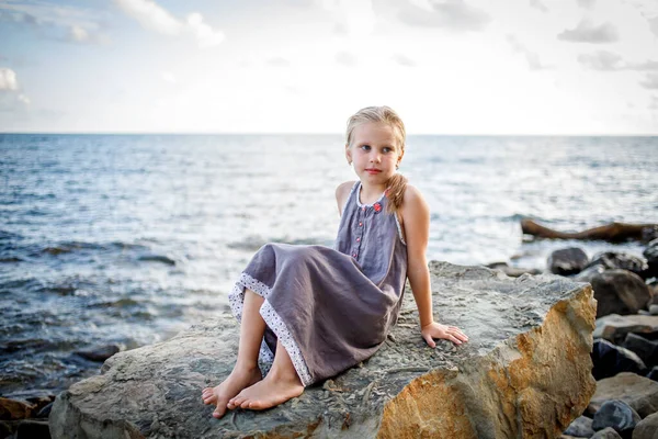 Blondýnka v slunečních šatech sedí na kameni na pobřeží. — Stock fotografie
