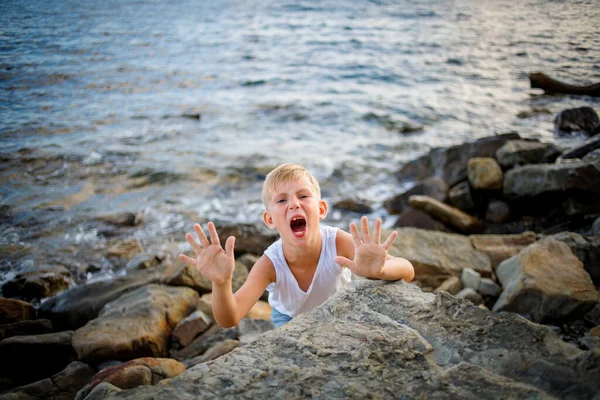Chlapec v bílém tričku a džínových kraťasech vykoukne zpoza velkého kamene na pobřeží. — Stock fotografie