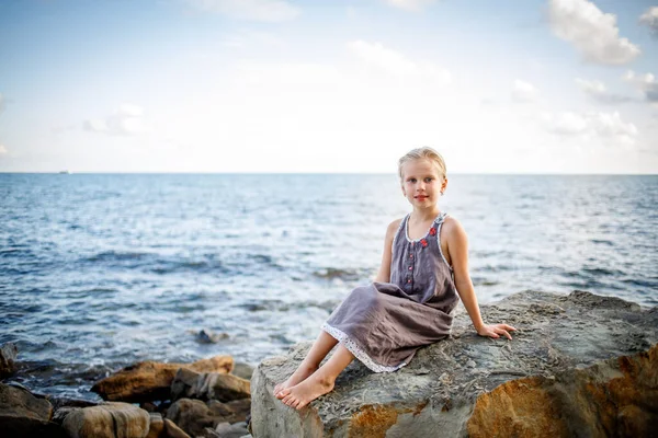 Blondýnka v slunečních šatech sedí na kameni na pobřeží. — Stock fotografie