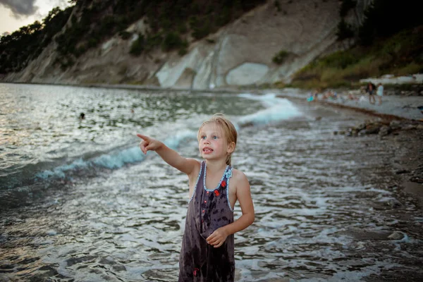 Ένα κορίτσι με μοβ φόρεμα στέκεται στη γραμμή του σερφ στο φόντο της θάλασσας και των βράχων. — Φωτογραφία Αρχείου