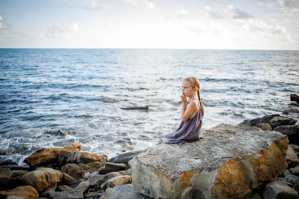 Uma menina loira em um vestido de verão senta-se em uma pedra na praia. — Fotografia de Stock