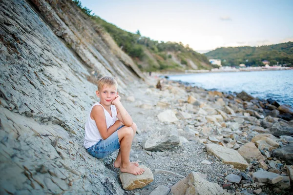 Chlapec v bílém tričku a džínových kraťasech sedí na skále u pobřeží — Stock fotografie