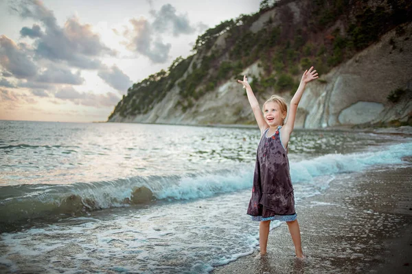 A girl in a purple sundress stands on the surf line against the background of the sea and rocks — Stock Photo, Image