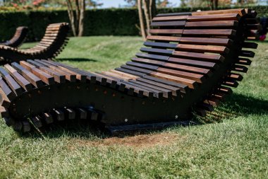 A curved chaise longue made of wooden slats stands on green grass in a city park.
