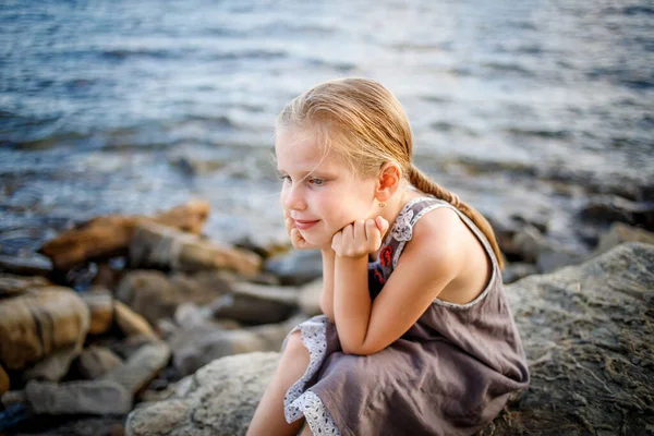 Une fille blonde en robe de soleil s'assoit sur une pierre au bord de la mer. — Photo
