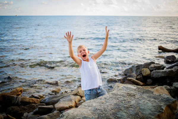 A boy in a white T-shirt and denim shorts jumps out from behind a large rock on the seashore. — Stock Photo, Image