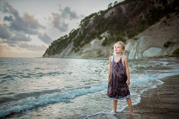 A girl in a purple sundress stands on the surf line against the background of the sea and rocks — Stock Photo, Image