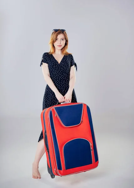 Brązowłosa dziewczyna w czarnej sukience stoi z dużą czerwono-niebieską walizką na szarym tle. — Zdjęcie stockowe