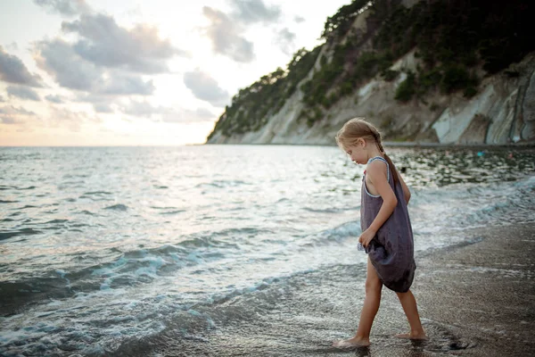 A girl in a purple sundress stands on the surf line against the background of the sea and rocks — Stock Photo, Image