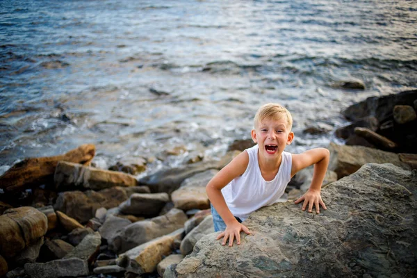 Chlapec v bílém tričku a džínových kraťasech vykoukne zpoza velkého kamene na pobřeží. — Stock fotografie