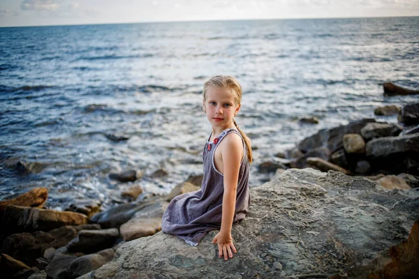 A blonde girl in a sundress sits on a stone on the seashore. — Stock Photo, Image