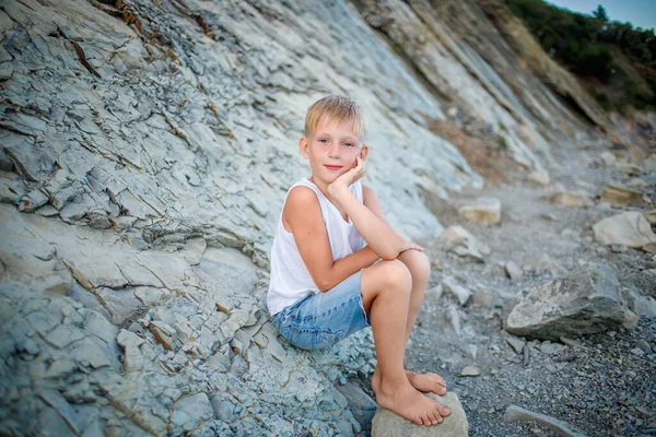 Un garçon en T-shirt blanc et short en denim est assis sur un rocher près du bord de la mer — Photo