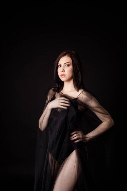 Siyah şifona sarılı siyah elbiseli esmer kız yarım tur önde duruyor.