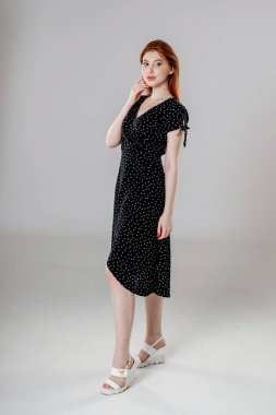 Siyah elbiseli siyah saçlı bir kız beyaz arka planda duruyor ve elleriyle saçlarını düzleştiriyor..