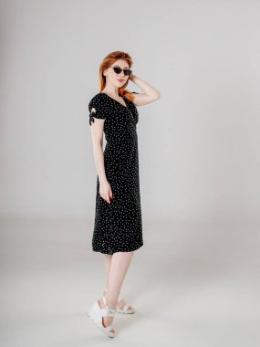 Siyah elbiseli siyah saçlı bir kız beyaz arka planda duruyor ve elleriyle saçlarını düzleştiriyor..