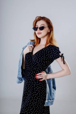 Siyah elbiseli siyah saçlı bir kız elinde kot ceketiyle beyaz bir arka planda duruyor..