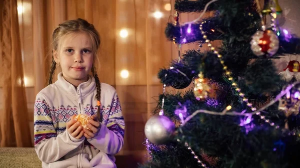 Une fille aux cheveux bruns nettoie une mandarine assise près d'un arbre de Noël décoré — Photo