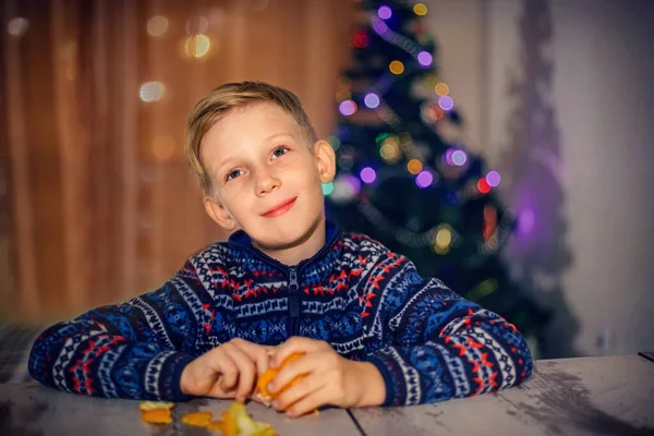 Un garçon aux cheveux bruns nettoie une mandarine assise près d'un arbre de Noël décoré — Photo