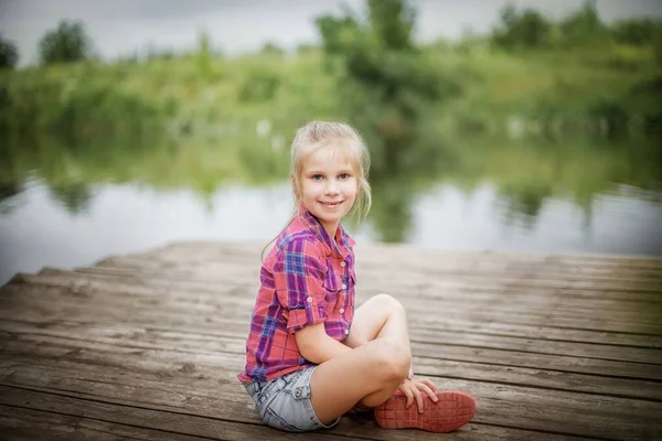 Une fille blonde est assise sur une jetée en bois au bord de la rivière. — Photo