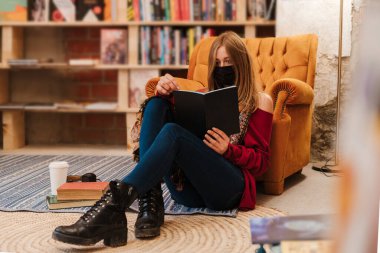 Maskeli genç sarışın kadın elinde kahveyle yerde oturuyor ve kitap okuyor.