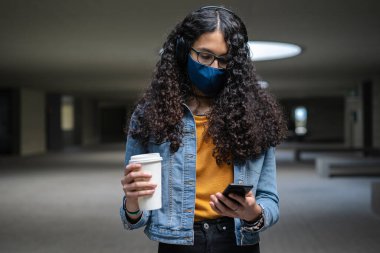 Yüz maskeli transseksüel kadın cep telefonuna bakıyor ve kahve içiyor.