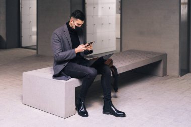 Yüzü maskeli bir iş adamı dizüstü bilgisayarıyla bankta otururken cep telefonuyla yazı yazıyor..