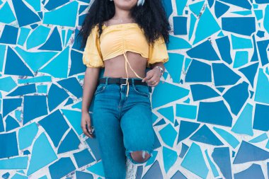 Sarı tişörtlü, mavi duvara yaslanmış kulaklıklarıyla müzik dinleyen tanınmamış Afro-Amerikan bir kadın..