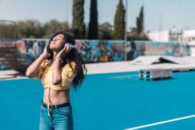 Sarı tişörtlü genç Afro-Amerikalı kız elinde kulaklıkla mutlu müzik dinliyor..