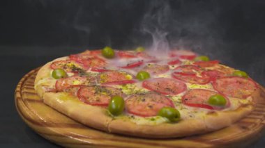 Eritilmiş peynirli taze pişmiş pizza, dilimlenmiş domates, yeşil zeytin ve ahşap servis tahtasındaki bitkiler, sıcak buhar çıkarıyor. Yüksek kalite 4k görüntü