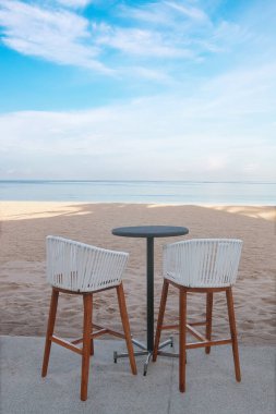 İki uzun bacak taburesi ve kumsalda bir siyah yuvarlak masa..