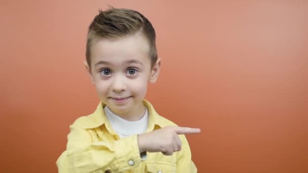 Pequeño niño emocional haciendo un gesto a un lado, mostrando espacio de copia sobre fondo naranja — Vídeos de Stock