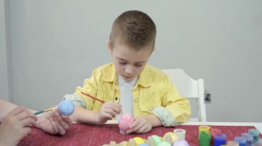 Paskalya yumurtalarını boyayan aile. Sarışın çocuk pembe renkli bir yumurtaya Paskalya desenleri çiziyor ve annesine gösteriyor. Mutlu Paskalyalar