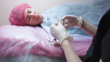 Dövme ustası işe hazırlanıyor. Güzellik uzmanı tek kullanımlık iğneyi açar ve dövme makinesine sokar. Arka plandaki müşteri güzellik uzmanının hazırlıklarını yakından takip ediyor.