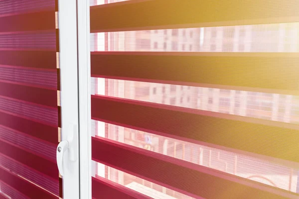 Evening sunlight enters the room through the blinds. Details of red fabric roller blinds on the plastic window in the room or kitchen.