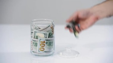 Adam parayı cam kavanoz bankasına yatırıyor. Evde para biriktirme kavramı. Cam bir kavanozda 100 dolarlık banknotlar. Ev finansmanı, yatırım, ekonomi