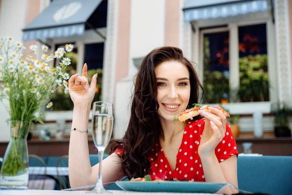 Brunette smiling female sits in outdoor cafeteria, with broad smile, has satisfied expression, feels relaxed and satisfied. Cute young woman with bruschetta in her hand rests in terrace cafe.