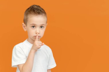 Sarışın, güzel, beyaz bir çocuk, sarı arka planda izole bir şekilde sessizlik işareti olarak işaret parmağıyla dudaklarını işaret etti. Okul öncesi çocuk portresi parmaklarıyla sessizlik hareketi yapıyor. Sansür kavramı.