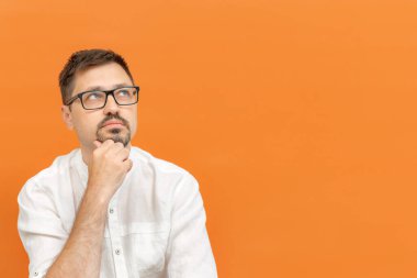 Gözlüklü yakışıklı beyaz adam düşünceli, meraklı ya da hayalperest, hayal kuruyor ve turuncu arka planda yer arıyor..