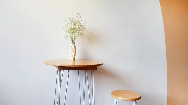 White flower in vase on table, wooden stool chair with vintage light on top with concrete wall and orange or brown pastel painted. Interior design and minimal space concept.