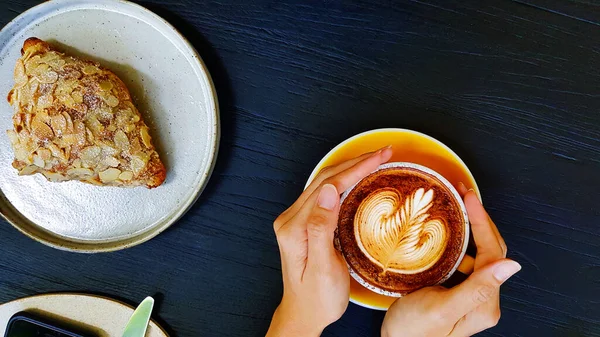 Kadının elinde badem kremalı latte kahve, bıçak, çatal ve telefon ile koyu mavi ahşap arka planda ya da fotokopi alanı olan bir masa. Sıcak kapuçino içiyorum..