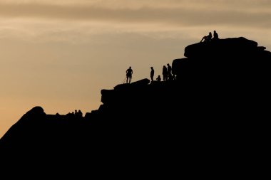 Stanage kenarındaki bir dağcıyı izleyen bir sürü insan silueti. Tırmanışçı fındık, ip ve karabina gibi aletlerle koşum takımı giyer..