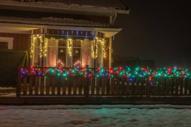 Ahşap kırmızı İsveç evinin penceresinde güzel bir manzara geleneksel şamdan ve ahşap teras çiti Noel ve yeni yıl için çok renkli aydınlatma, evin önünde ıslak kar.